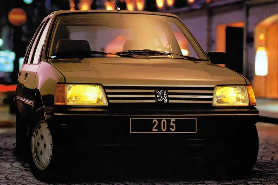 Peugeot 205 éclairée la nuit, phares allumés, sur rue pavée.