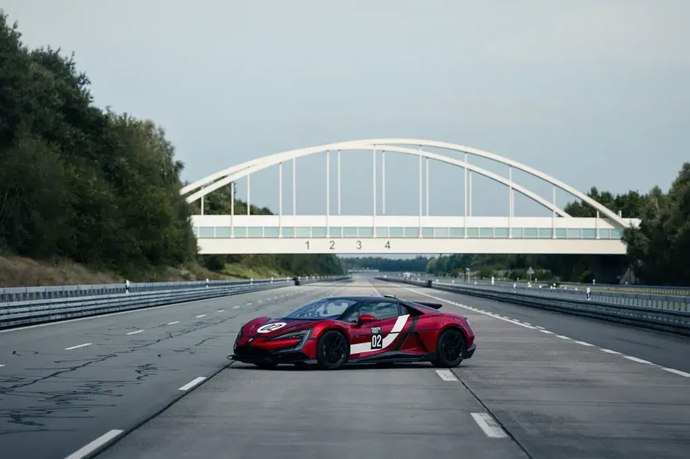 Rode McLaren sportwagen op testcircuit onder witte brug, bosrijke omgeving op achtergrond.