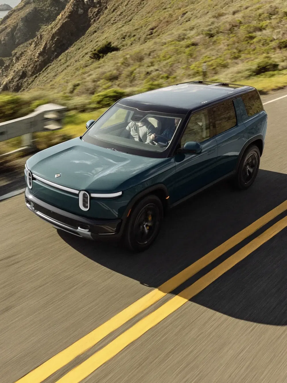 Rivian R1S électrique roulant sur une route sinueuse, entourée de montagnes verdoyantes.