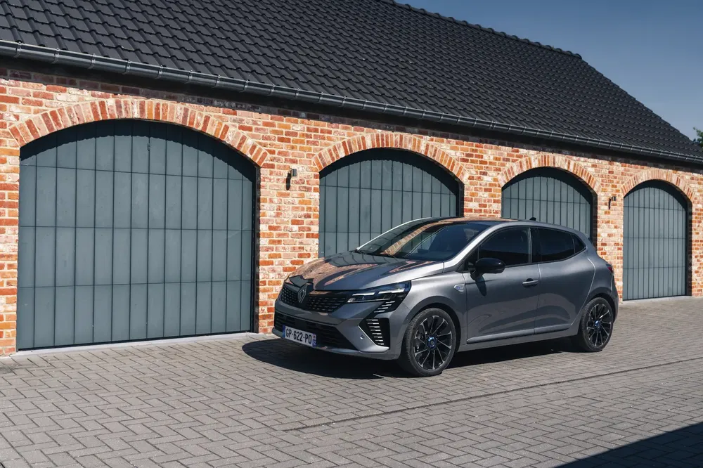 Grijze Renault Clio geparkeerd voor een bakstenen gebouw met blauwe deuren.