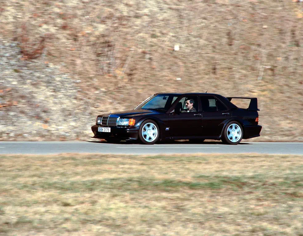 Mercedes 190E 2.3-16 roulant sur route, aileron arrière proéminent, arrière-plan de paysage aride.