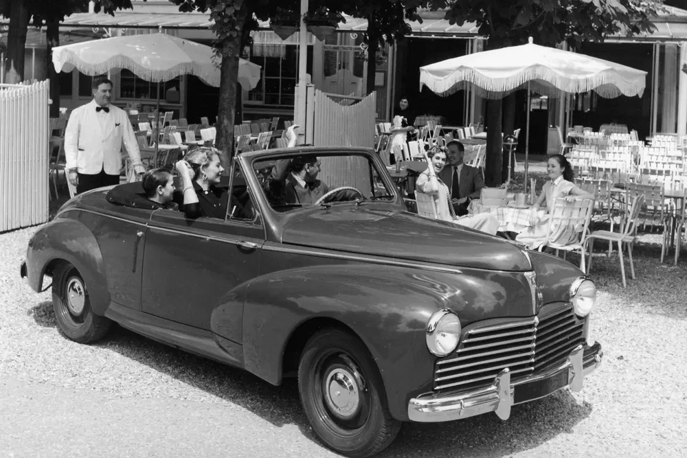 Peugeot 203 décapotable des années 1950 avec passagers, garé devant un café en terrasse.