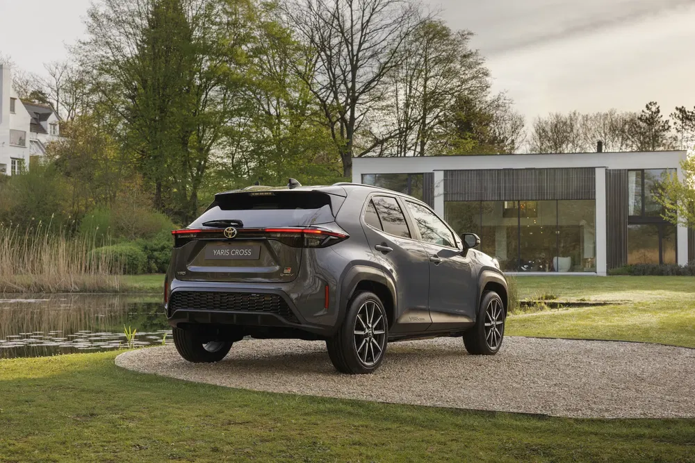 Grijze Toyota Yaris Cross voor moderne woning bij vijver, omringd door groen.