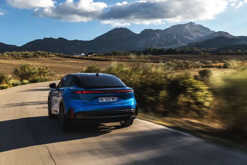Alpine électrique bleu sur route de campagne avec montagnes en arrière-plan, ciel partiellement nuageux.