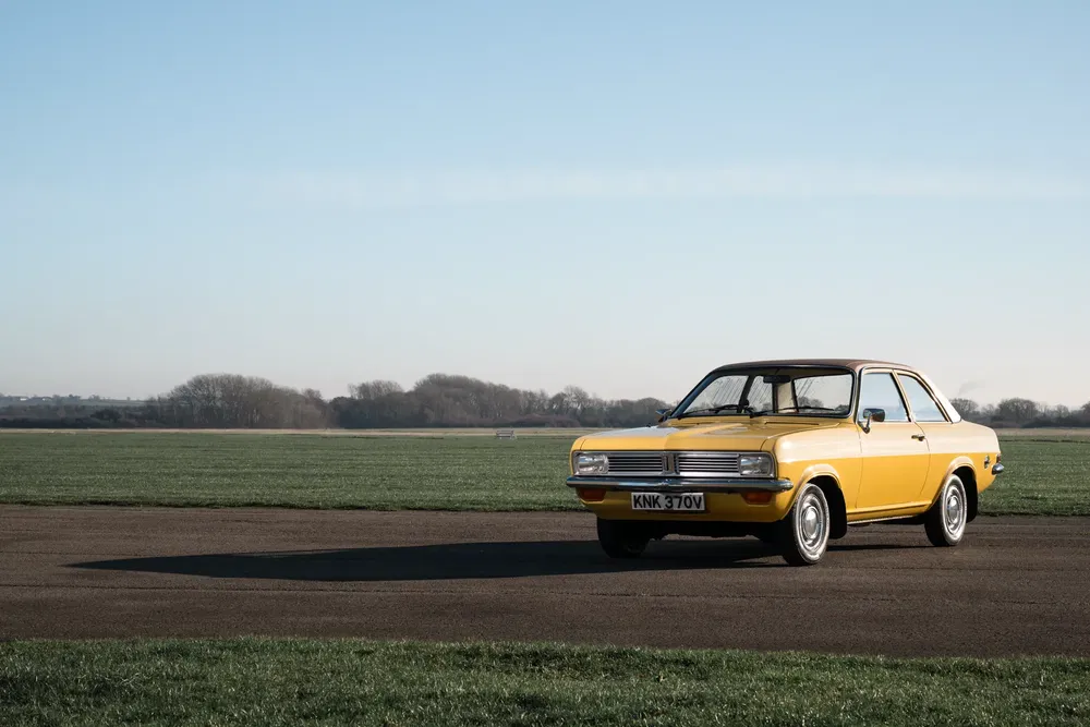 Vauxhall vintage jaune sur route, arrière-plan champ verdoyant et ciel bleu.