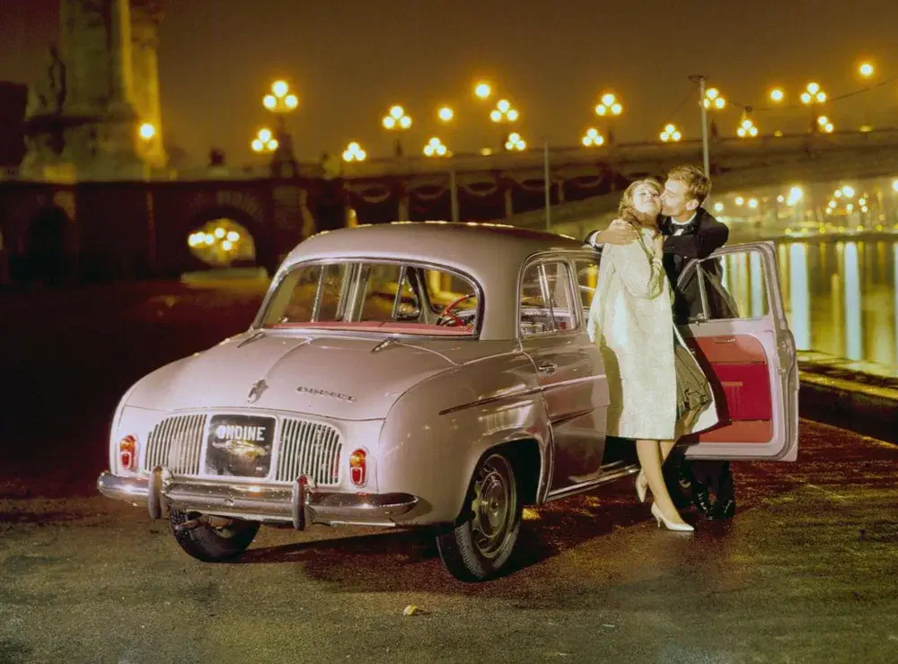 Couple élégant posant près d'une Renault Dauphine rose, sur fond de lumières nocturnes.