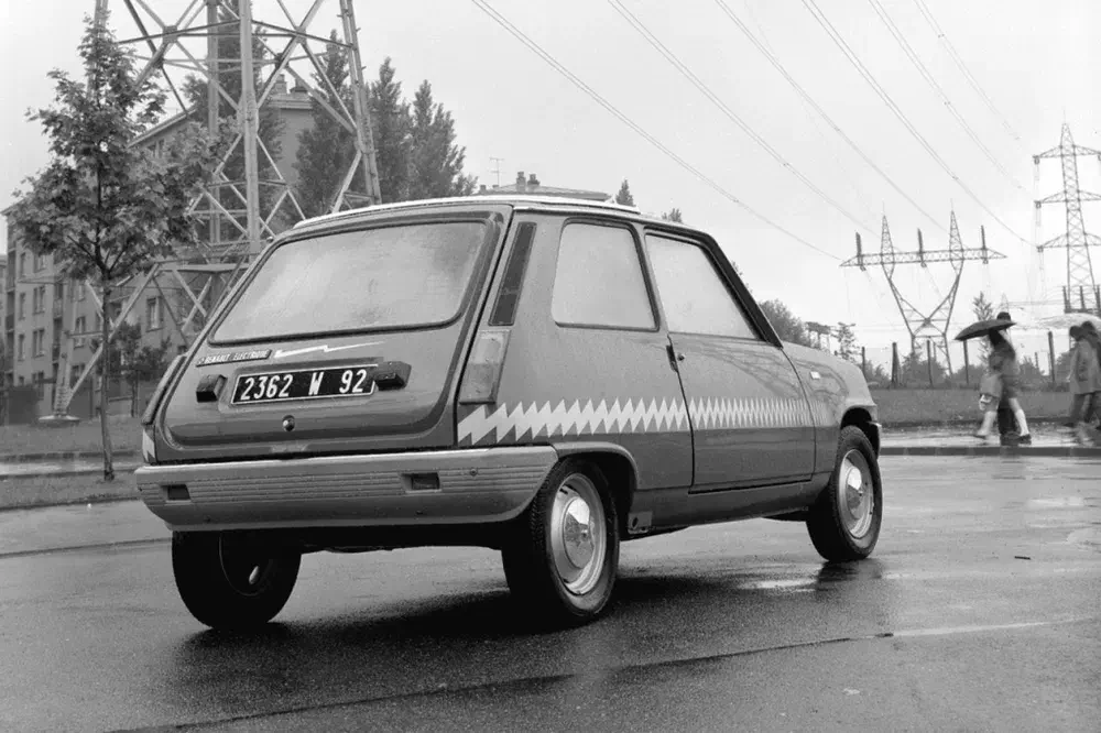 Renault 5 classique roulant sous la pluie, arrière distinctif, ambiance urbaine en arrière-plan.