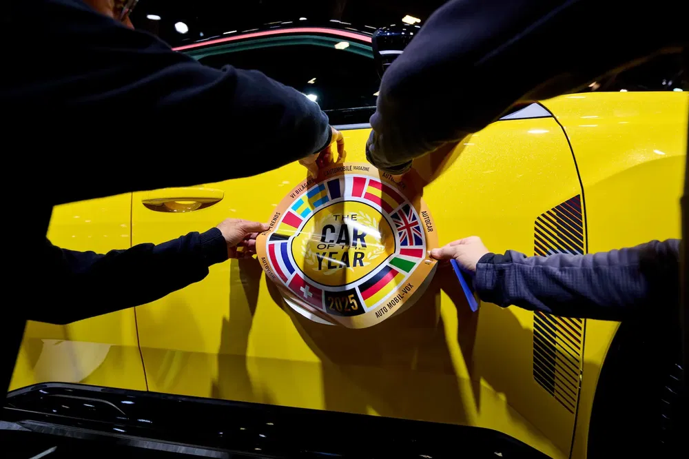 Twee handen plaatsen 'Car of the Year'-sticker op gele auto, vlaggen zichtbaar.