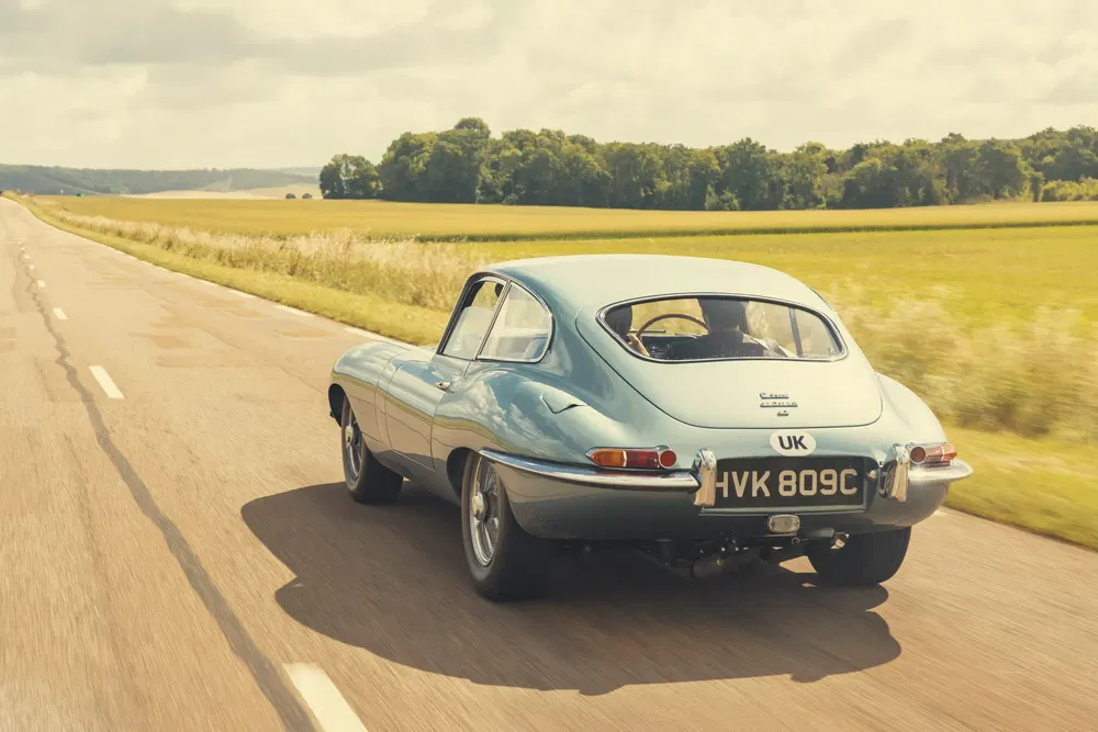 Jaguar E-Type klassieker rijdt op zonnige landweg, groen veld op de achtergrond.