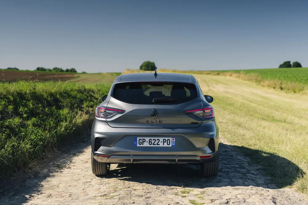 Grijze Renault Clio op een geplaveid pad omgeven door groene velden, achteraanzicht.
