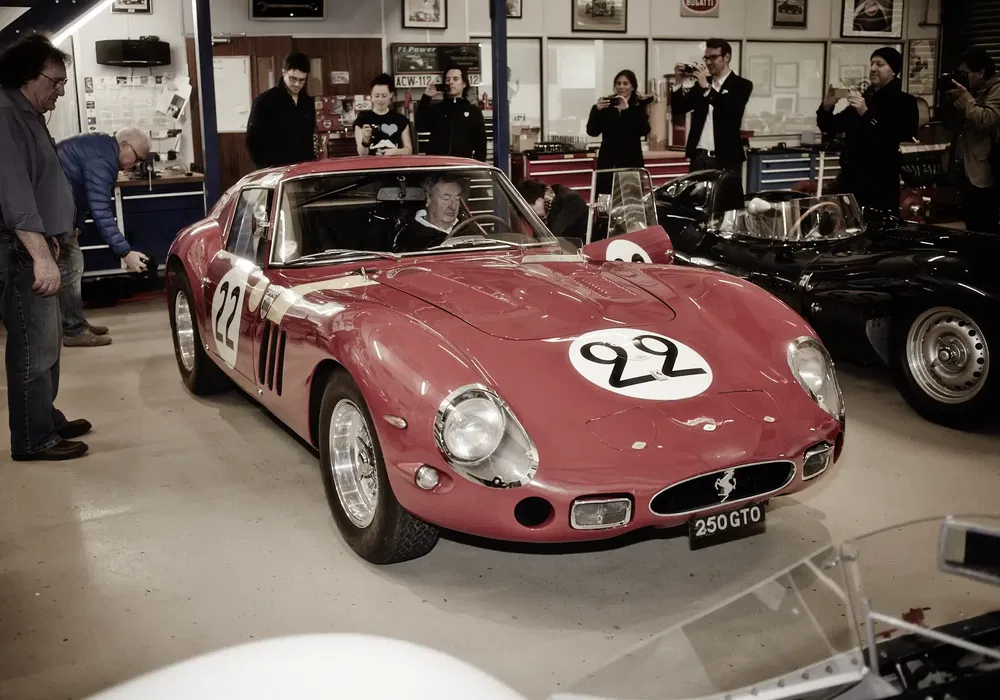 Ferrari 250 GTO rouge dans un garage, entourée de spectateurs et photographiée intensément.