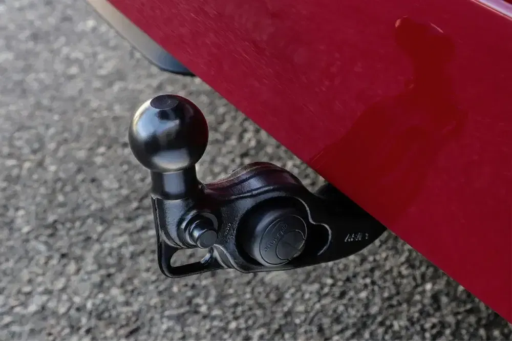 Crochet d'attelage noir fixé à une voiture rouge sur asphalte.