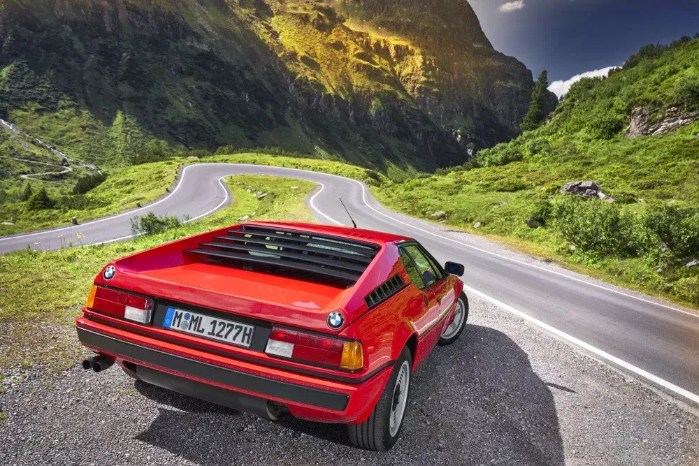 BMW M1 rouge sur route de montagne sinueuse, paysage verdoyant alpin.