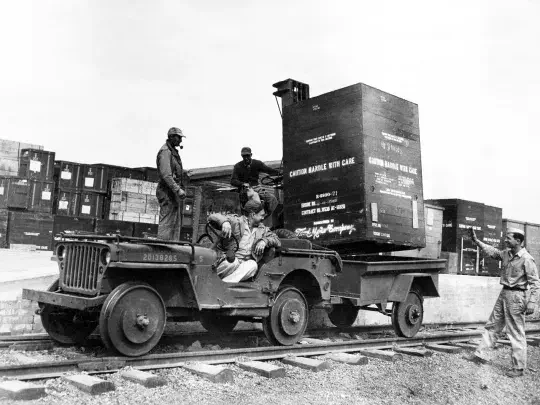 Militaire Jeep vervoert grote kist op spoorplatform, soldaten in actie.