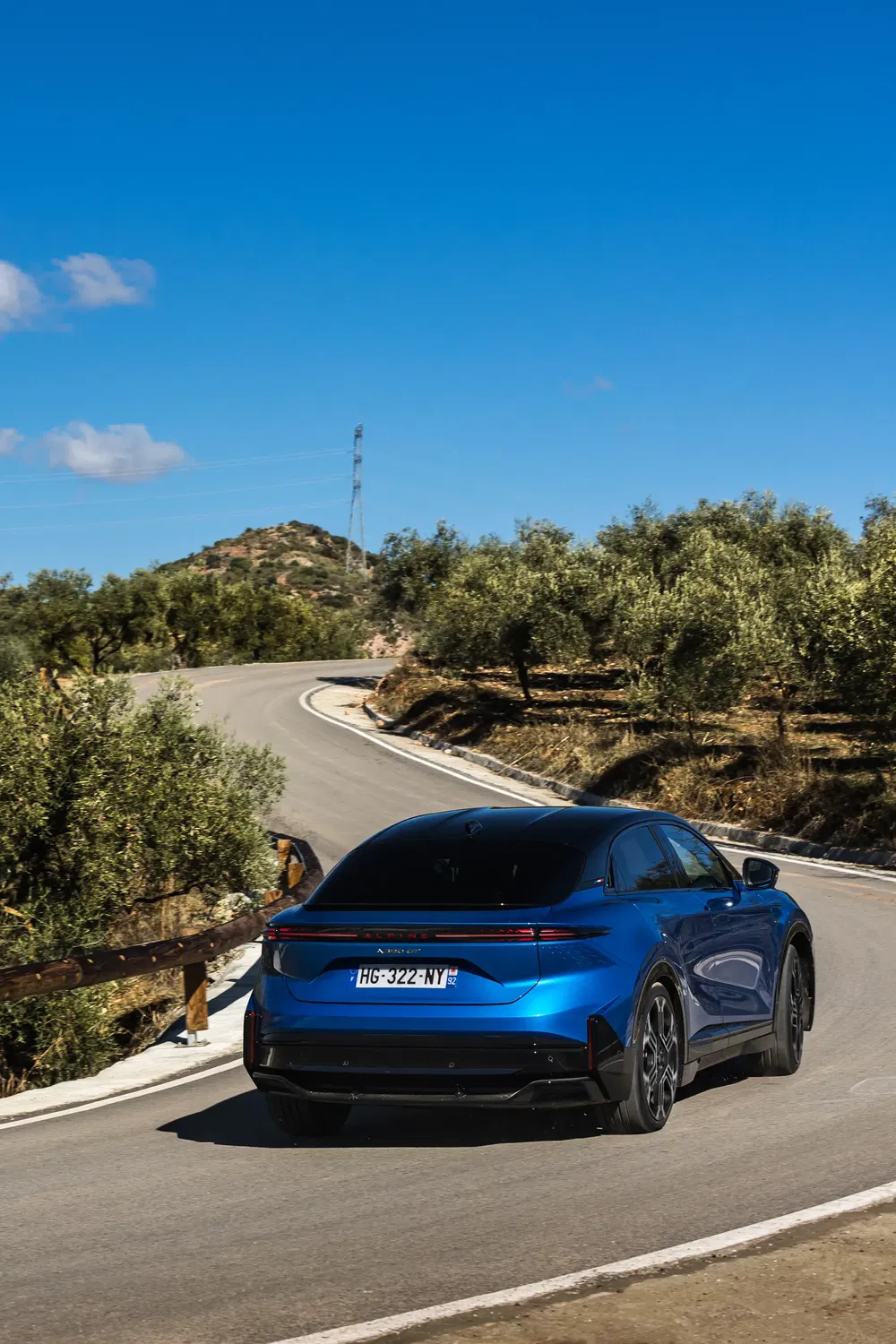 Alpine A290 bleu sur route sinueuse entourée d'arbres, ciel bleu dégagé.