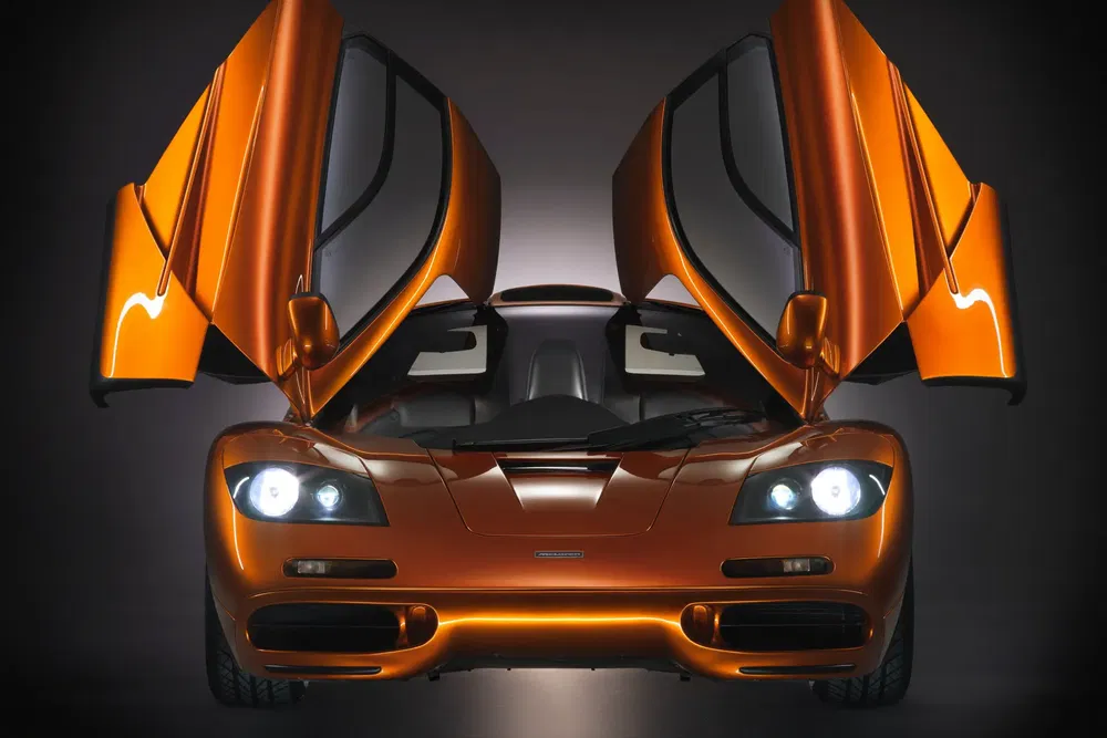Voiture McLaren F1 orange avec portes papillon ouvertes, vue frontale dynamique.