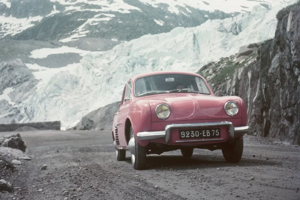 Voiture rétro rouge sur route de montagne, paysage enneigé en arrière-plan, immatriculation française.