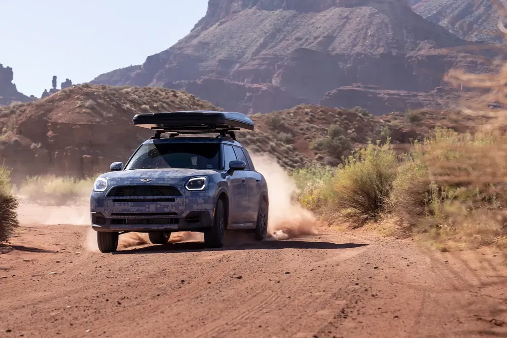 Mini Countryman 2024 met camouflage rijdt door een bergachtig woestijnlandschap.