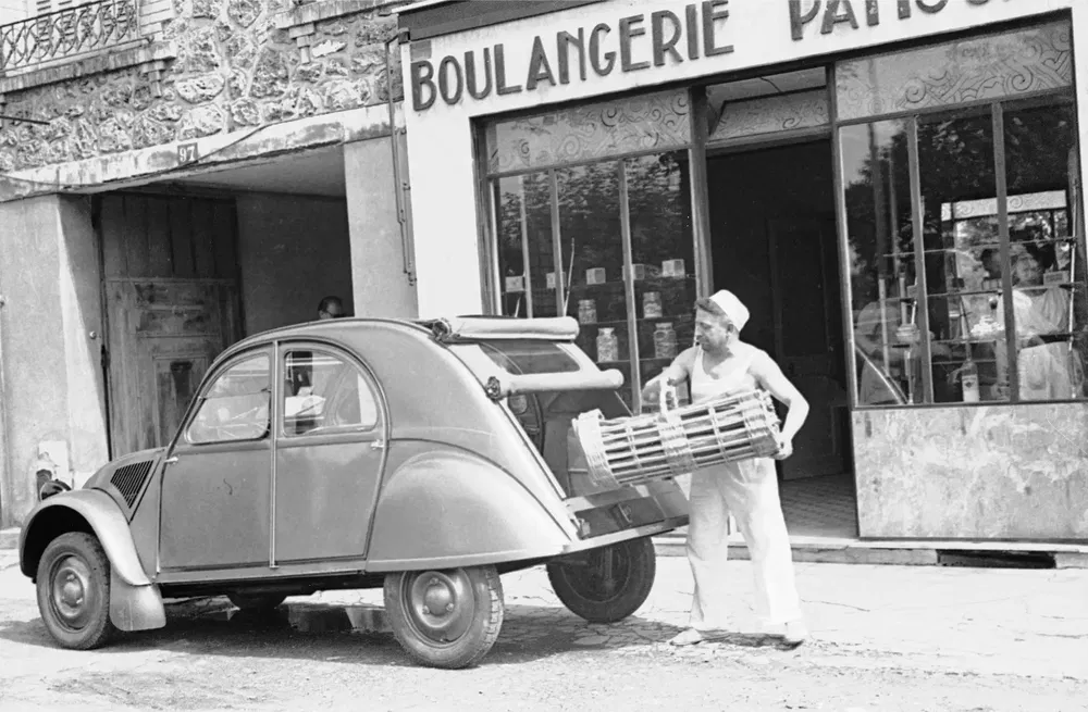 Citroën 2CV devant une boulangerie avec un boulanger transportant des paniers à pain.