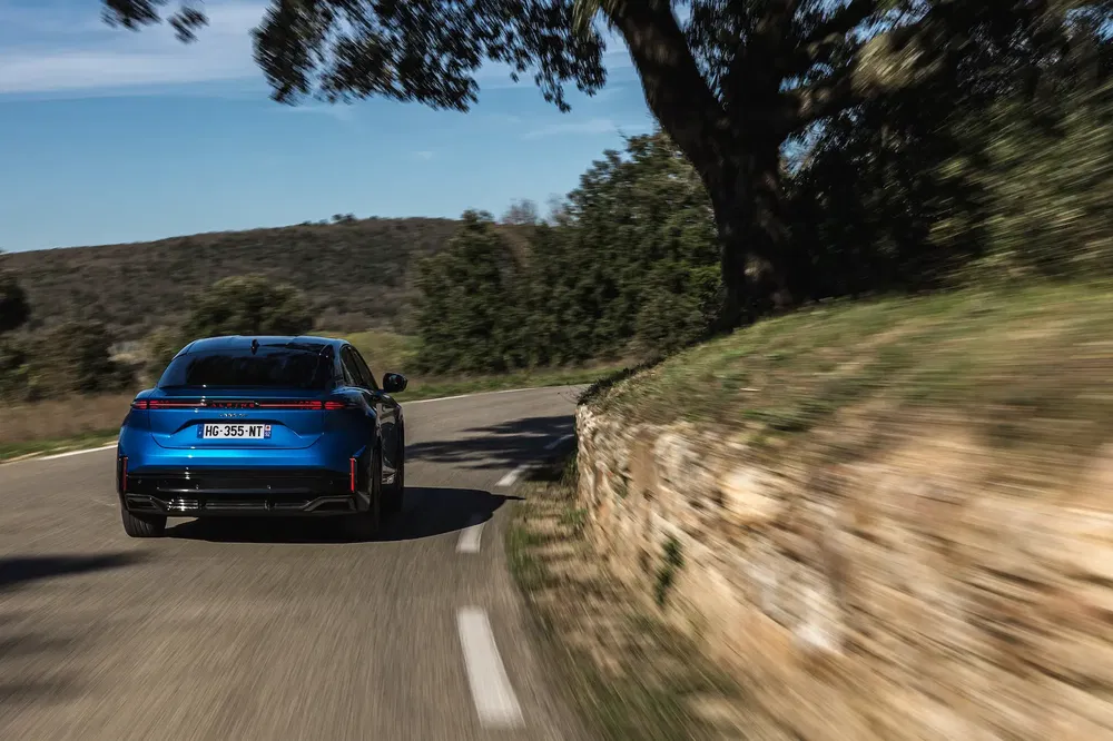 Blauwe elektrische Peugeot 508 rijdt op bochtige landelijke weg, heldere blauwe lucht.