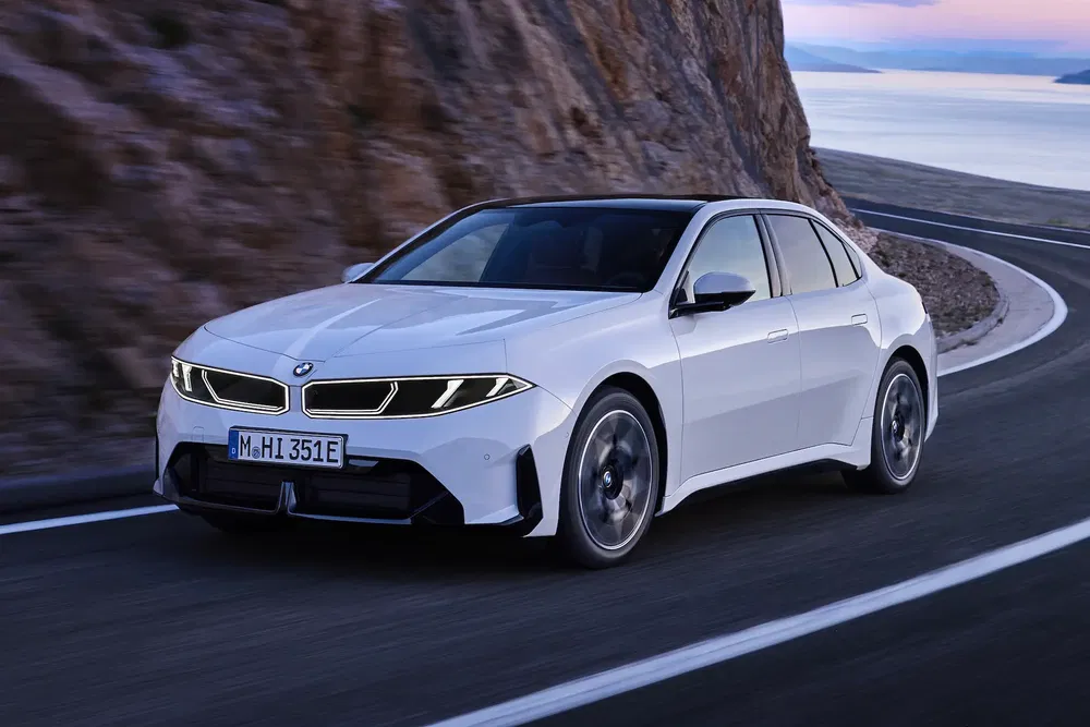 BMW électrique blanche sur route côtière avec falaise, au coucher du soleil.