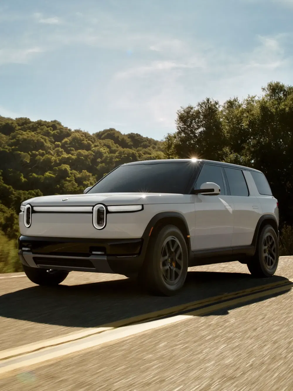 SUV Rivian R1S blanc roulant sur route bordée d'arbres, design moderne et écologique.