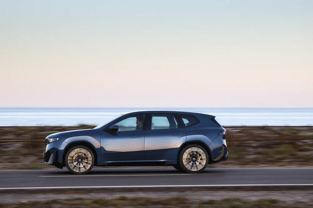Blauwe BMW iX rijdt langs kustweg bij zonsondergang, met zee op de achtergrond.