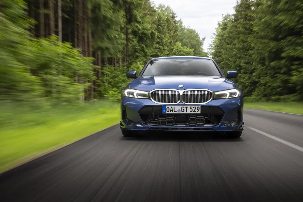 BMW Série 3 bleue roulant sur route forestière, avant dynamique en mouvement.