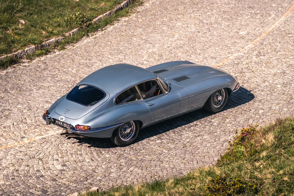 Klassieke zilvergrijze Jaguar E-Type op kronkelige geplaveide weg.
