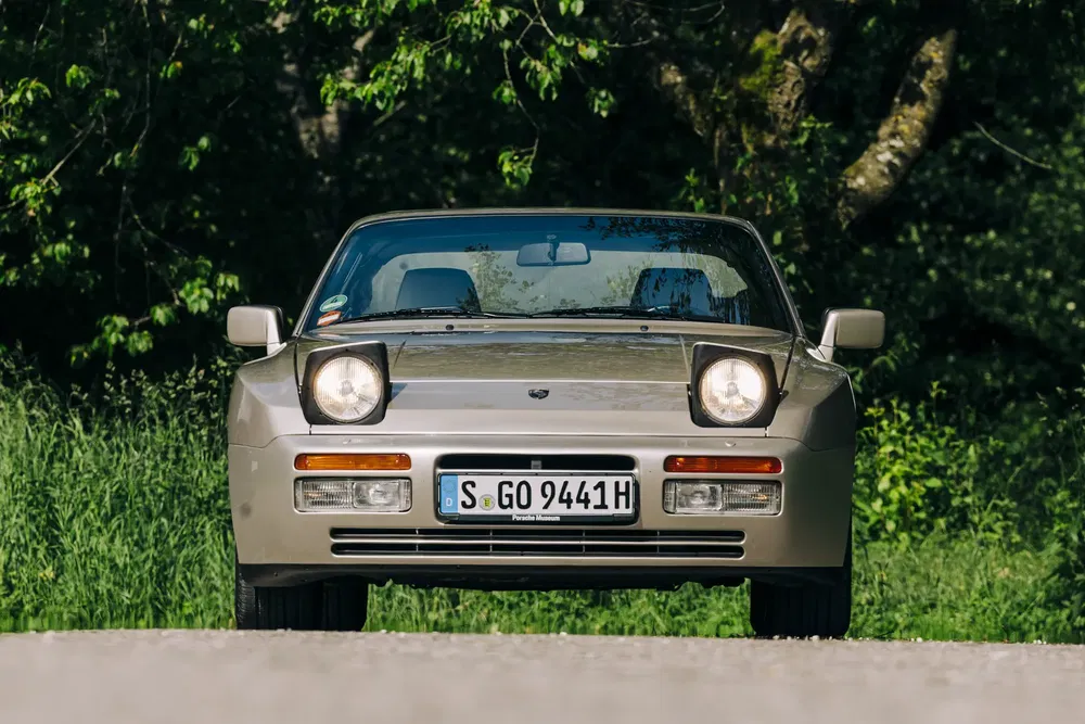 Porsche 944 argenté sur une route bordée d'arbres verdoyants, vue de face.