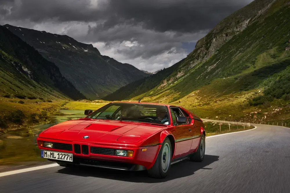 BMW M1 rouge sur route de montagne, paysage montagneux dramatique en arrière-plan.