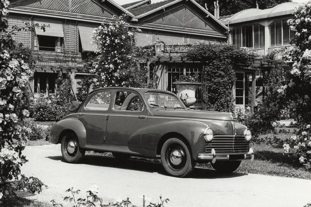 Peugeot 203 classique garée devant une maison vintage, entourée de rosiers en fleurs.
