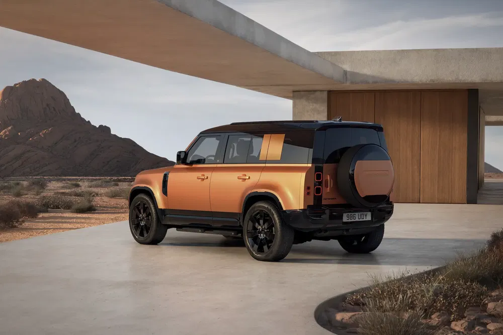Land Rover Defender orange stationné dans un désert devant une maison moderne.