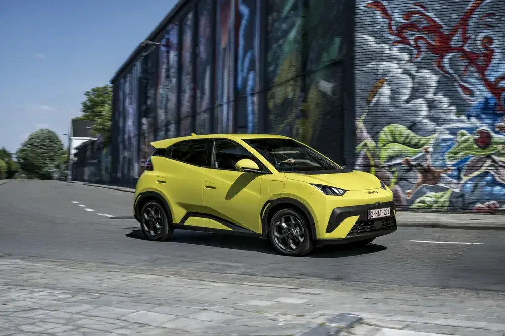 Voiture électrique jaune BYD Atto 3 passant devant un mur coloré de graffiti en ville.