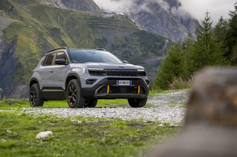 Jeep Avenger grise sur chemin montagneux avec arrière-plan de montagnes et forêts verdoyantes.