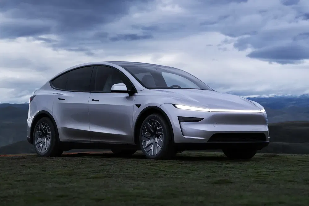 Tesla Model Y argenté sur colline herbeuse, ciel nuageux en arrière-plan.