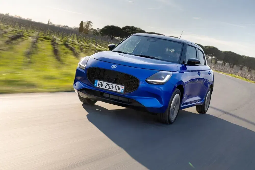 Suzuki Swift bleue sur route de campagne avec arrière-plan champêtre et ciel dégagé.