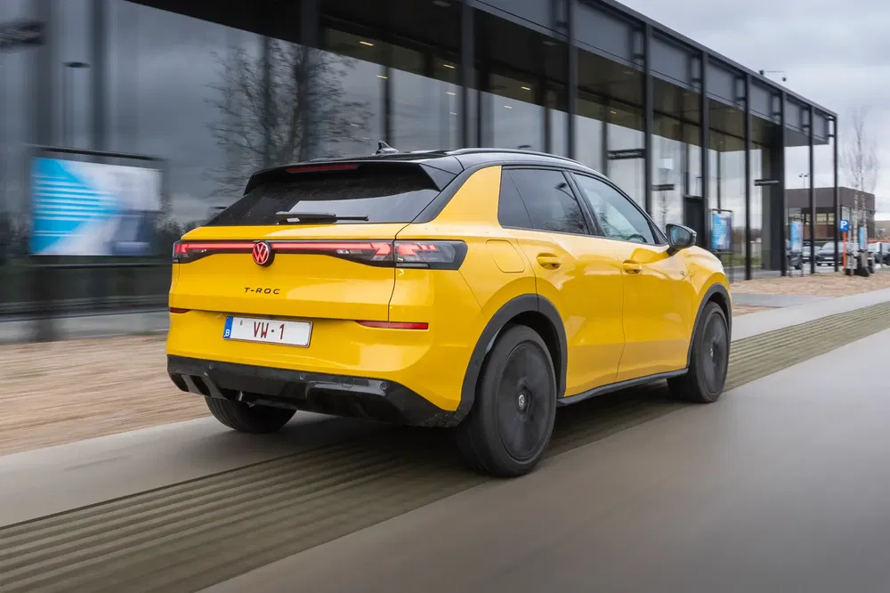 Volkswagen T-Roc jaune roulant devant un bâtiment moderne par temps nuageux.