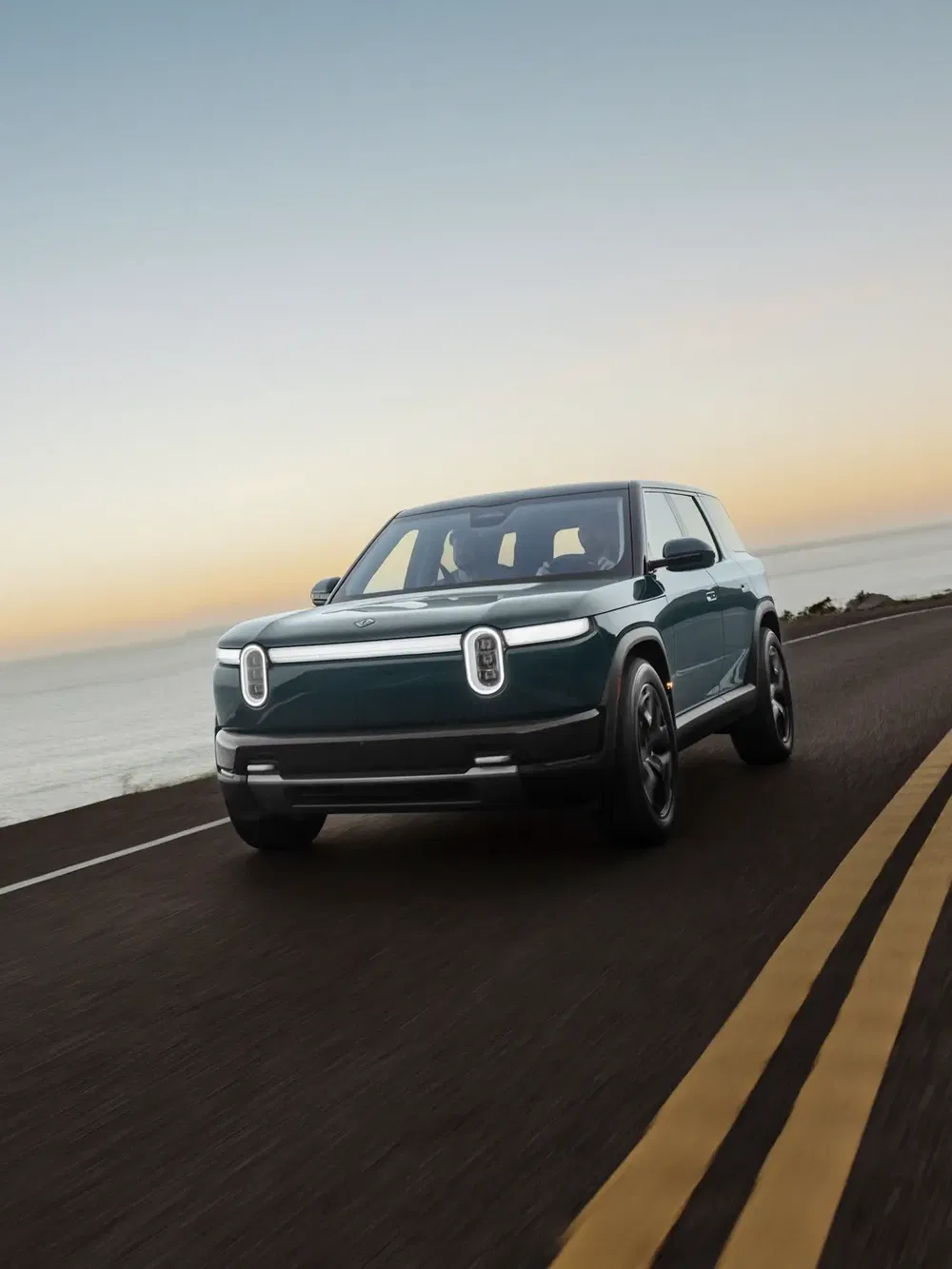 Rivian R1S électrique roulant sur autoroute côtière au coucher de soleil.