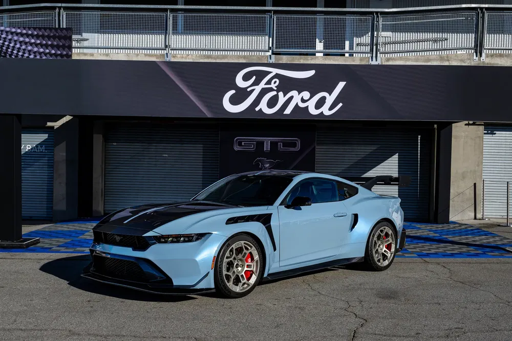 Ford Mustang GTD bleu avec aileron, stationnée devant une enseigne Ford sur circuit.