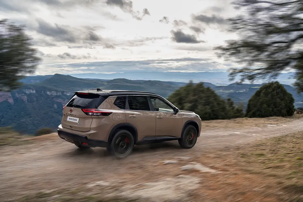 Beige Nissan X-Trail rijdt op een zandweg met panoramisch bergzicht.