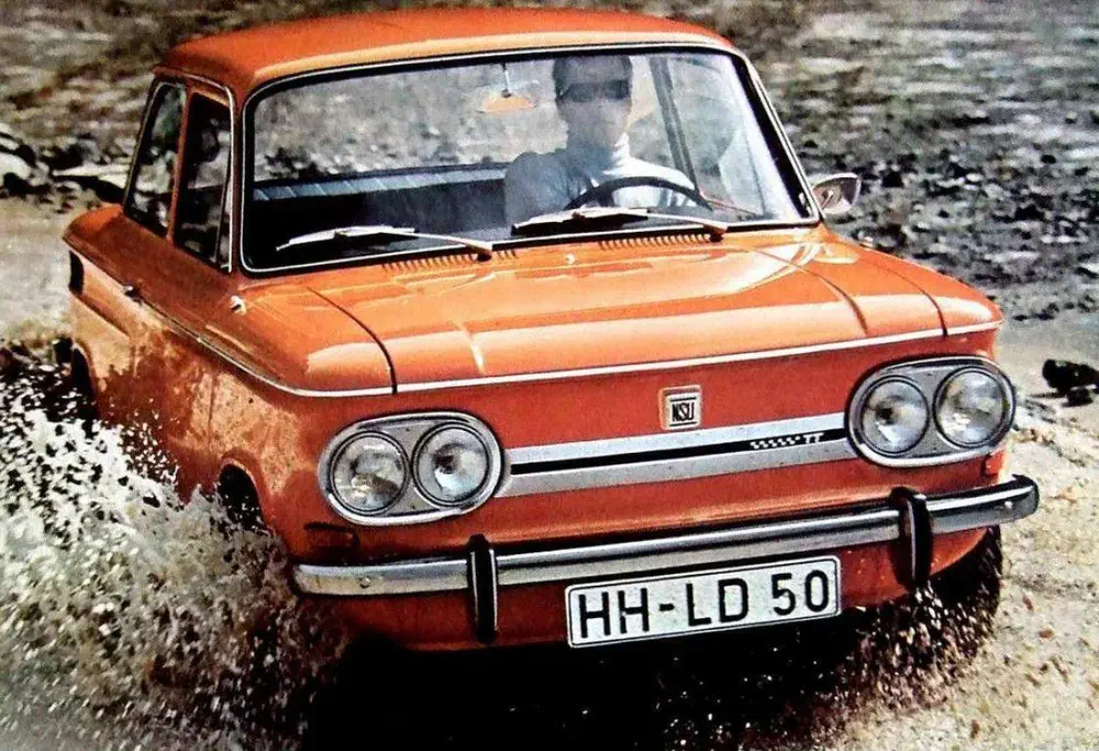 {"fr":"Voiture classique NSU orange traversant une flaque d'eau avec élégance.","nl":"Klassieke oranje NSU-auto rijdt stijlvol door een plas water."}