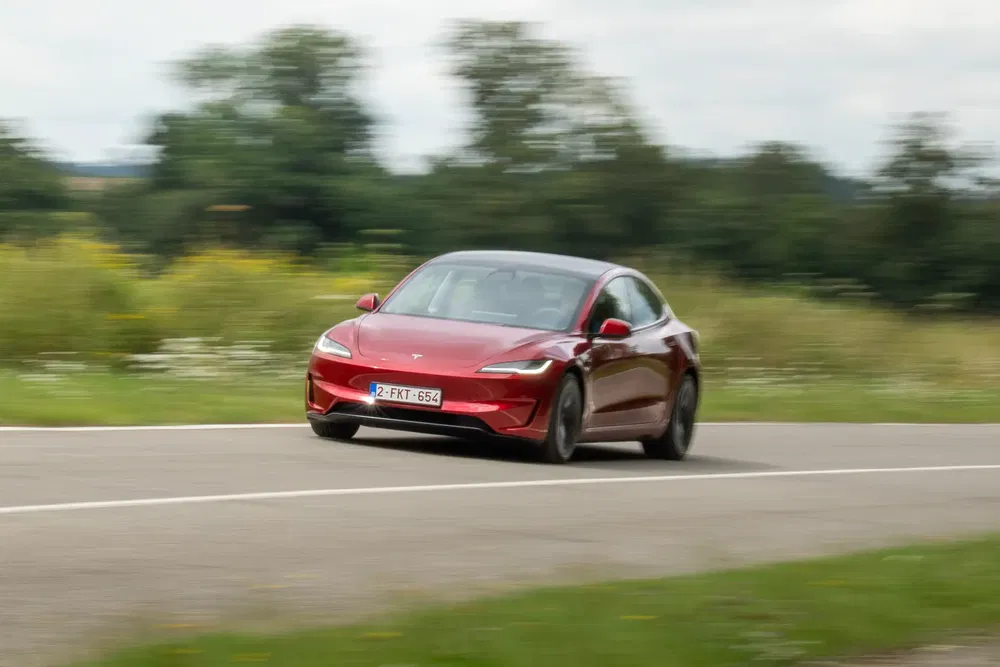 Rode Tesla Model 3 rijdt op plattelandsweg met bomen op de achtergrond.