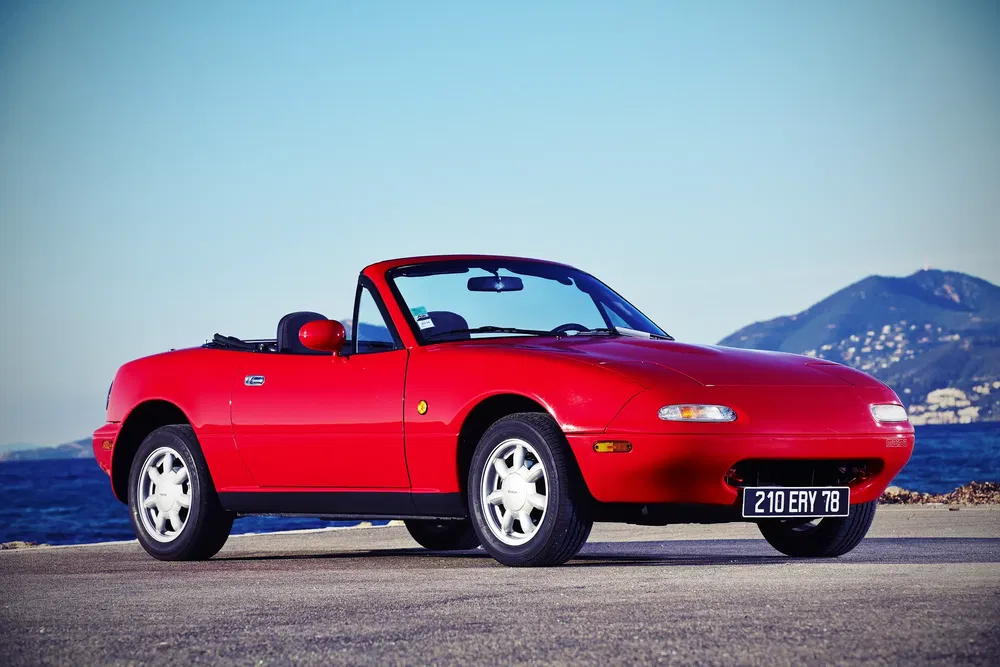 Mazda MX-5 rouge décapotable au bord de la mer avec vue sur les montagnes.