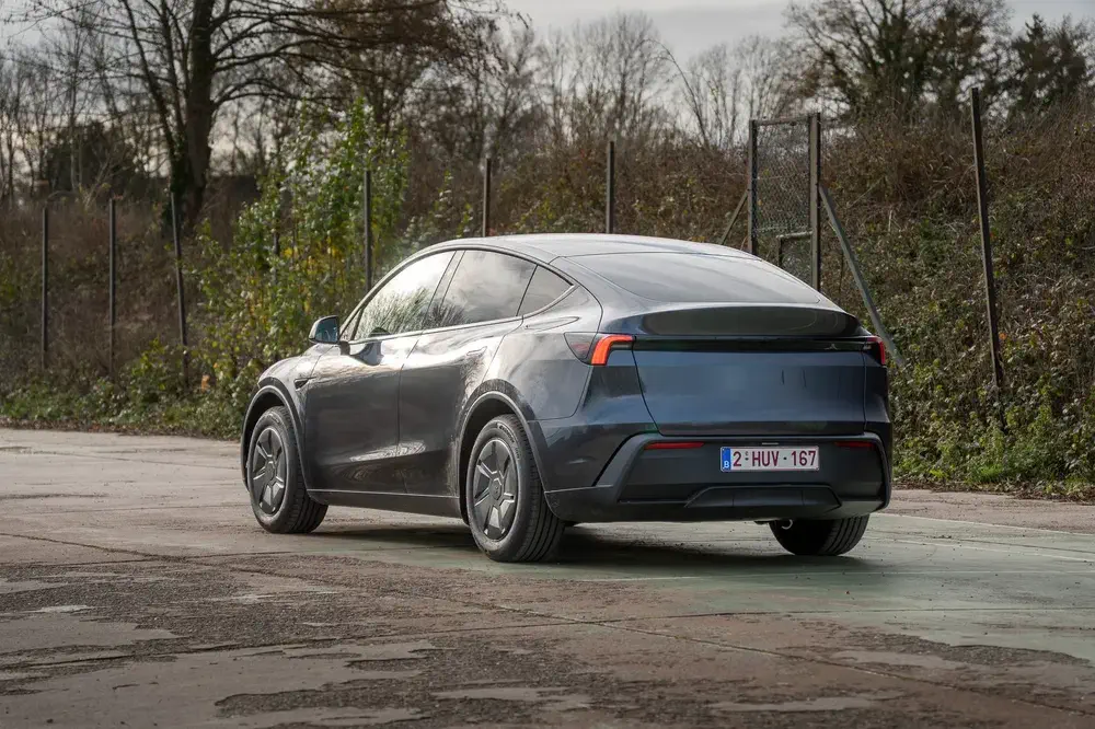 Tesla Model Y gris sur une route pavée, entouré d'arbres et de clôtures métalliques.