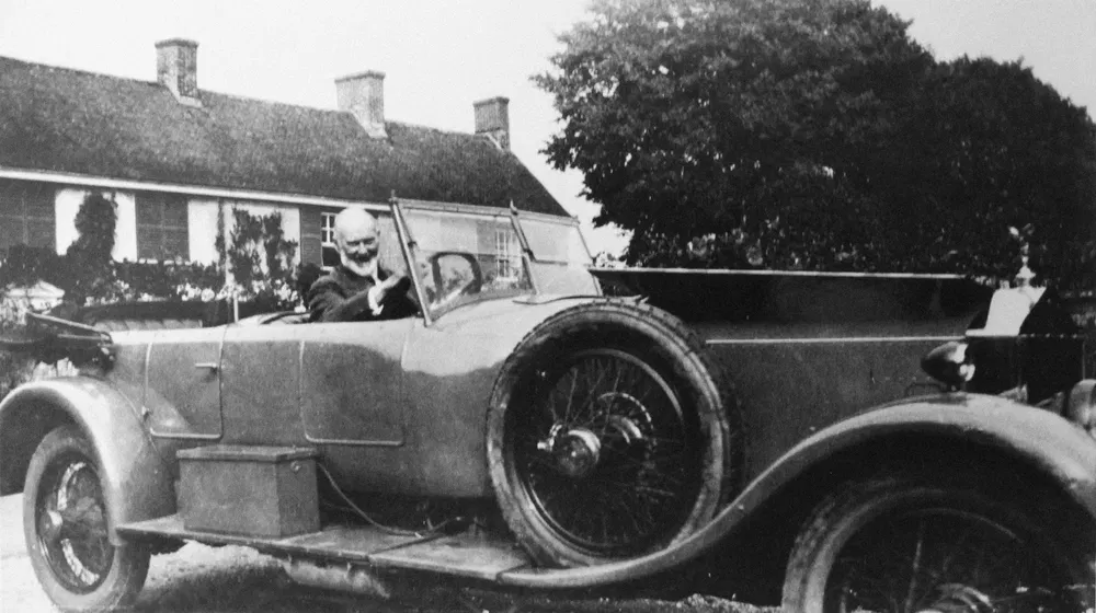 Voiture classique ancienne avec conducteur souriant devant une maison rurale.