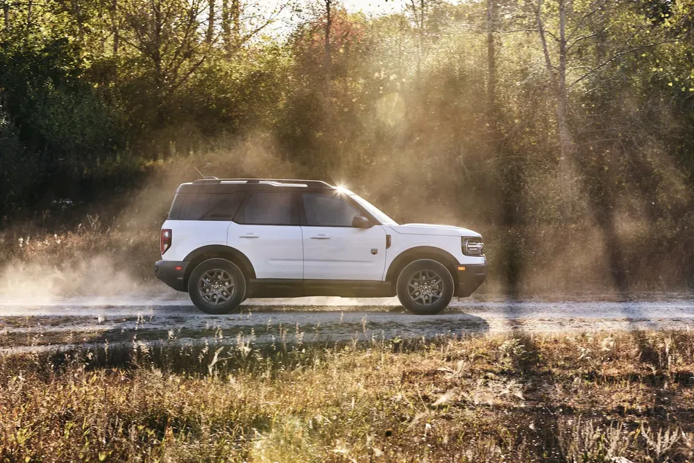 SUV Ford Bronco Sport blanc roulant sur chemin poussiéreux en forêt sous soleil couchant.