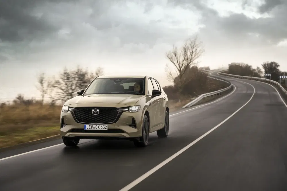 Mazda CX-60 beige roulant sur route sinueuse sous ciel nuageux.