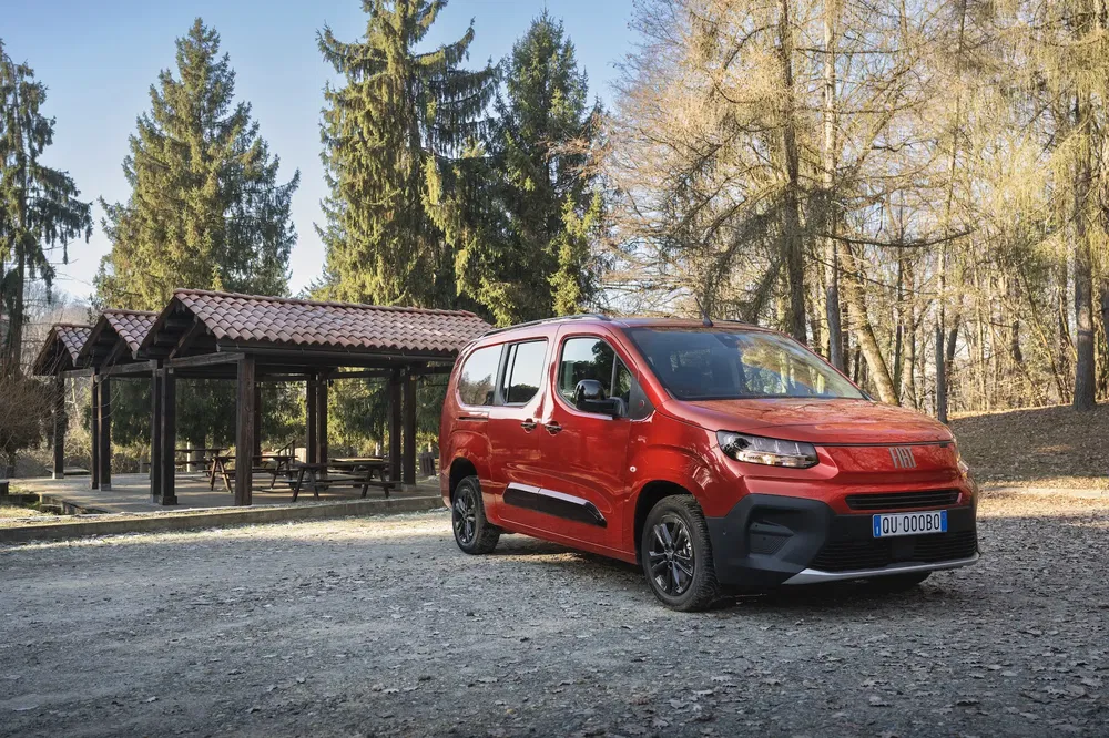 Rode Fiat Doblo geparkeerd bij een houten schuilplaats in een bosrijk park.