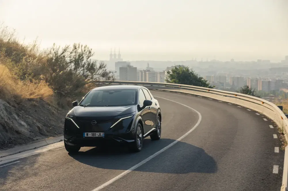Zwarte Nissan Ariya rijdt op een kronkelige weg met uitzicht op een verlichte stad.